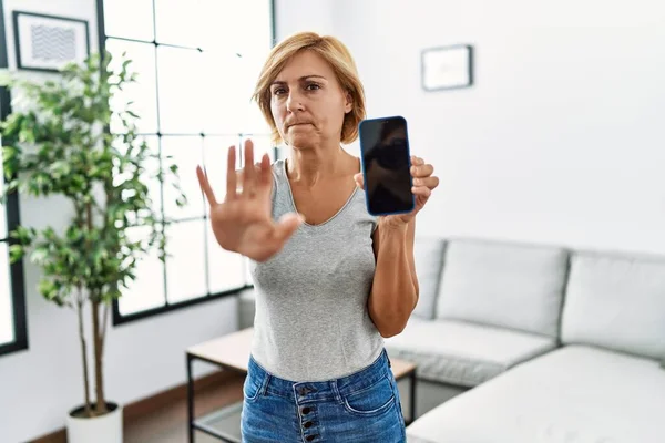Middle age blonde woman working holding smartphone showing screen with open hand doing stop sign with serious and confident expression, defense gesture