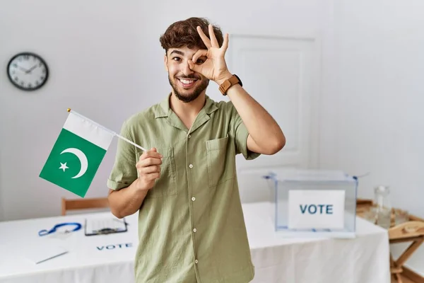 Young arab man at political campaign election holding pakistan flag smiling happy doing ok sign with hand on eye looking through fingers