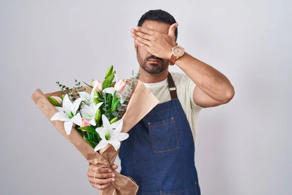Hispanic man with beard working as florist covering eyes with hand, looking serious and sad. sightless, hiding and rejection concept