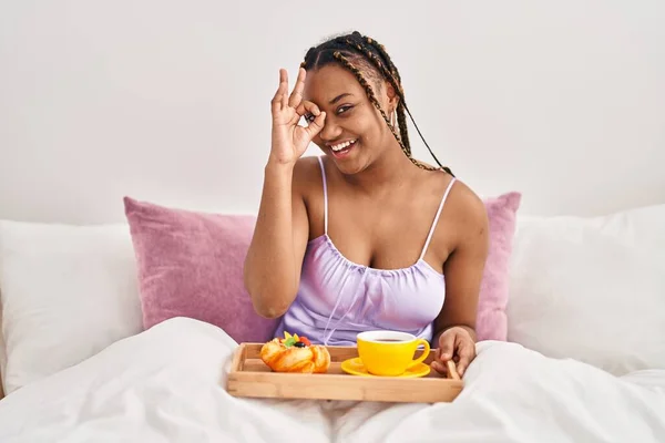 African american woman with braids holding tray with breakfast food in the bed smiling happy doing ok sign with hand on eye looking through fingers