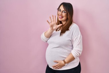 Pregnant woman standing over pink background showing and pointing up with fingers number five while smiling confident and happy. 