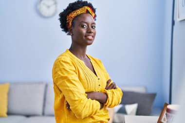 Genç Afrikalı Amerikalı kadın gülümsüyor. Kendine güveni tam. Kollarını kavuşturup evde duruyor.