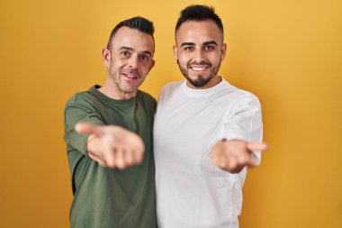 Homosexual couple standing over yellow background smiling cheerful offering palm hand giving assistance and acceptance. 