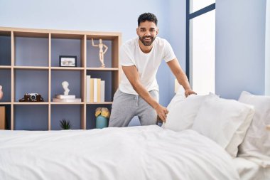 Young hispanic man make bed standing at bedroom