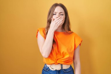 Caucasian woman standing over yellow background bored yawning tired covering mouth with hand. restless and sleepiness. 