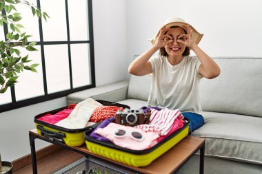 Güzel, orta yaşlı İspanyol bir kadın valize yazlık giysiler yerleştiriyor. Dürbün gibi jest yapıyor. Dilini çıkarıyor, gözleri parmaklarından bakıyor. çılgın ifade. 