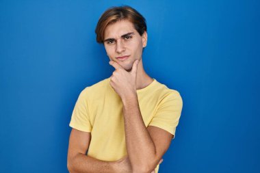 Young man standing over blue background looking confident at the camera with smile with crossed arms and hand raised on chin. thinking positive. 