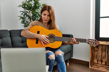 Orta yaşlı beyaz bir kadın online gitar dersi alıyor. Evdeki kanepede oturuyor..