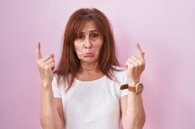 Middle age woman standing over pink background pointing up looking sad and upset, indicating direction with fingers, unhappy and depressed. 