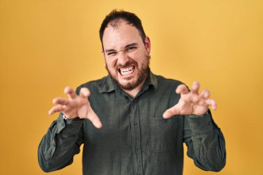Plus size hispanic man with beard standing over yellow background smiling funny doing claw gesture as cat, aggressive and sexy expression 