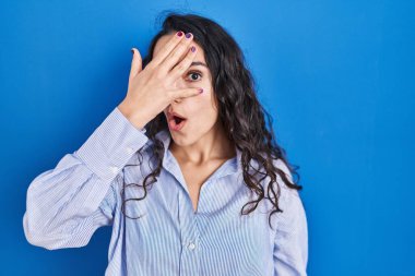 Genç esmer kadın mavi arka planda dikiliyor. Şoka girip yüzünü ve gözlerini kapatıyor. Utanmış bir ifadeyle parmaklarından bakıyor.. 
