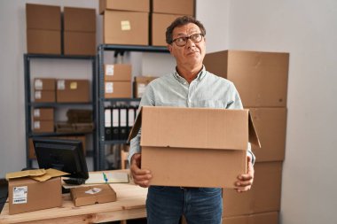 Senior man working at small business ecommerce holding carboard bx smiling looking to the side and staring away thinking. 