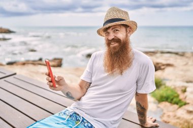 Genç kızıl saçlı turist akıllı telefon kullanıyor sahildeki bankta oturuyor..