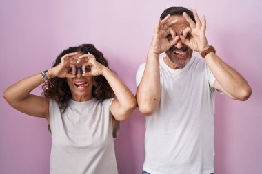 Orta yaşlı İspanyol çift pembe arka planda birlikte dürbün gibi jest yapıyorlar, gözleri parmaklarından bakıyor. çılgın ifade. 