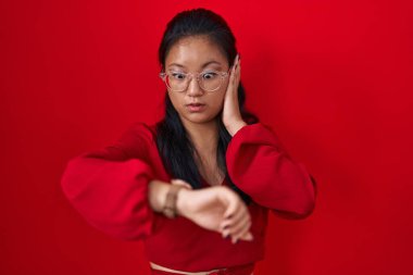Asian young woman standing over red background looking at the watch time worried, afraid of getting late 