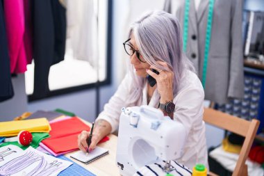 Orta yaşlı, kır saçlı kadın terzi terzide akıllı telefon yazısıyla konuşuyor.