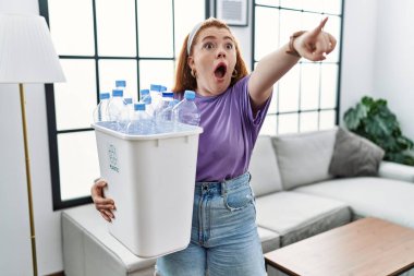 Elinde geri dönüşüm sepetini tutan genç kızıl saçlı bir kadın. Elinde parmakları önde olan plastik şişeler var. Şaşırmış, ağzı açık, ön tarafta bir şey var. 