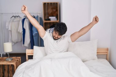 Young hispanic man waking up stretching arms at bedroom