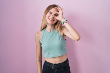 Sarışın, beyaz bir kadın pembe arka planda duruyor. El hareketleriyle gülümsüyor, gözleriyle mutlu yüzüyle parmaklarının arasından bakıyor.. 