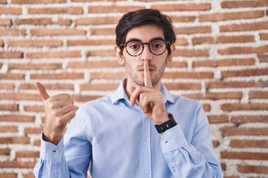 Young hispanic man standing over brick wall background asking to be quiet with finger on lips pointing with hand to the side. silence and secret concept. 