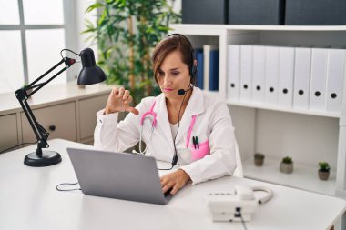 Middle age doctor woman working wearing headset with angry face, negative sign showing dislike with thumbs down, rejection concept 