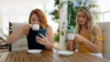 Two women using smartphones and drinking coffee sitting on table at home terrace