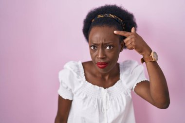 African woman with curly hair standing over pink background pointing unhappy to pimple on forehead, ugly infection of blackhead. acne and skin problem 