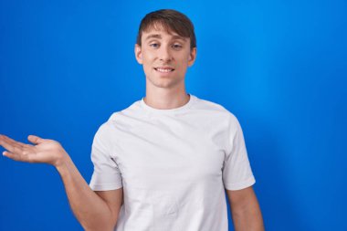 Caucasian blond man standing over blue background smiling cheerful presenting and pointing with palm of hand looking at the camera. 