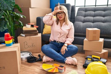 Young hispanic woman moving to a new home sitting on the floor making fun of people with fingers on forehead doing loser gesture mocking and insulting. 