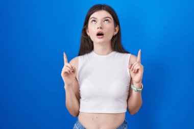Mavi arka plan üzerinde duran genç beyaz kadın şaşırmış ve şaşırmış bir şekilde yukarı bakıyor ve parmaklarıyla işaret ediyor ve kollarını kaldırıyor.. 