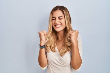 İzole edilmiş arka planda duran genç sarışın kadın başarı için heyecanlanıyor kolları havada ve gözleri kapalı zaferi kutlarken gülümsüyor. Kazanan konsept. 