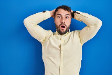 Handsome hispanic man standing over blue background crazy and scared with hands on head, afraid and surprised of shock with open mouth 