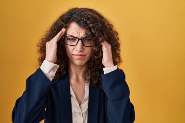 Hispanic woman with curly hair standing over yellow background with hand on head for pain in head because stress. suffering migraine. 