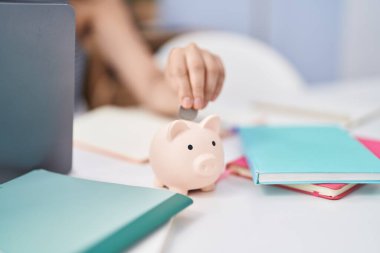 Young hispanic girl inserting coin on piggy bank at home