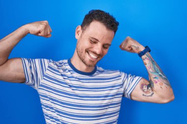 Mavi arka planda duran İspanyol genç adam gururla gülümseyen kol kaslarını gösteriyor. fitness konsepti. 