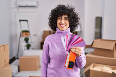 Yeni ev duvarının rengini seçen kıvırcık saçlı genç esmer kadın. Pozitif ve mutlu görünüyor. Kendine güvenen bir gülümsemeyle dişlerini gösteriyor. 