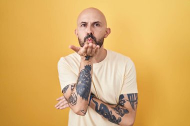 Sarı arka planda dikilmiş İspanyol dövmeli bir adam kameraya bakıp havada öpücük savuruyor ve güzel ve seksi oluyor. aşk ifadesi. 