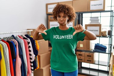 Young african american woman wearing volunteer t shirt at donations stand pointing down looking sad and upset, indicating direction with fingers, unhappy and depressed. 