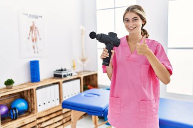 Genç fizyoterapist kadın sağlık merkezinde masaj tabancası tutuyor mutlu ve pozitif gülümsüyor, başparmağıyla mükemmel ve onay işareti yapıyor. 