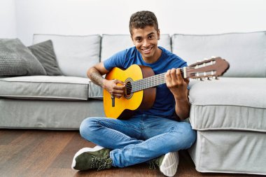 Gitar çalan genç İspanyol adam evde yerde oturuyor.