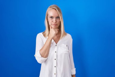 Mavi arka planda duran beyaz bir kadın kameraya kollarını kavuşturup gülümseyerek ve çenesini kaldırarak bakıyor. Olumlu düşünmek. 