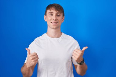 Caucasian blond man standing over blue background success sign doing positive gesture with hand, thumbs up smiling and happy. cheerful expression and winner gesture. 