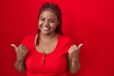 African american woman with braided hair standing over red background success sign doing positive gesture with hand, thumbs up smiling and happy. cheerful expression and winner gesture. 
