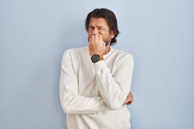 Mavi arka planda sıradan süveter giyen yakışıklı orta yaşlı bir adam. Ağzı tırnak yiyen elleri yüzünden gergin ve stresli görünüyor. anksiyete. 