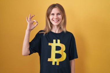 Bitcoin tişörtü giyen sarışın beyaz kadın gülümsüyor ve el ve parmaklarıyla tamam işareti yapıyor. başarılı ifade. 