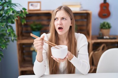Young caucasian woman eating asian food using chopsticks angry and mad screaming frustrated and furious, shouting with anger. rage and aggressive concept. 