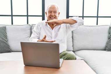 Üst düzey bir adam evinde dizüstü bilgisayar kullanıyor. Kanepede oturuyor. Elleri büyük ve büyük bir işaret, ölçü işareti gösteriyor. Kameraya bakıp gülümsüyor. ölçüm kavramı. 