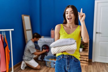 Young brunette woman holding folded laundry surprised with an idea or question pointing finger with happy face, number one 