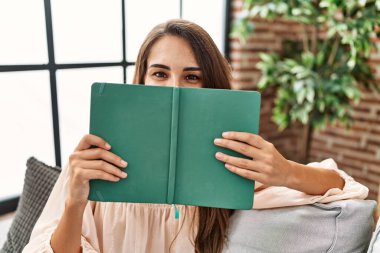 Young hispanic woman covering mouth with book at home