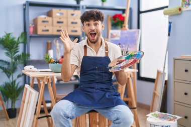 Arab man with beard painter sitting at art studio holding palette celebrating victory with happy smile and winner expression with raised hands 